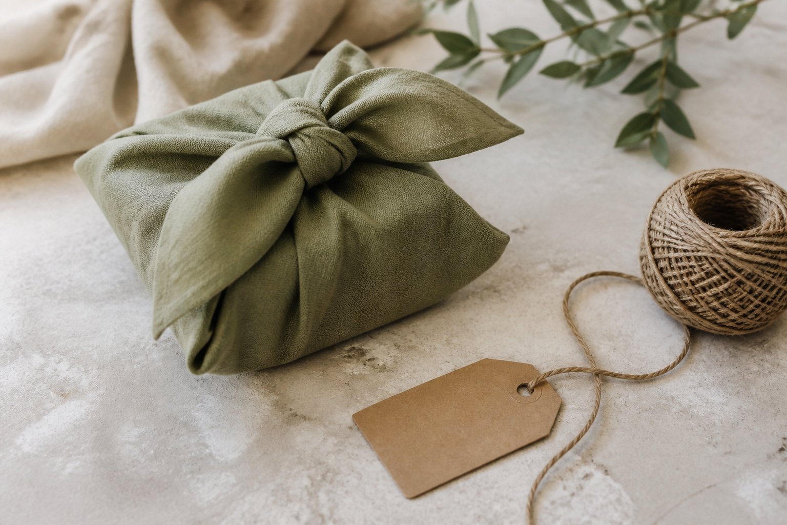 Cadeau emballé dans un carré de tissu noué, posé à côté d’une étiquette en papier kraft et d’une pelote de ficelle